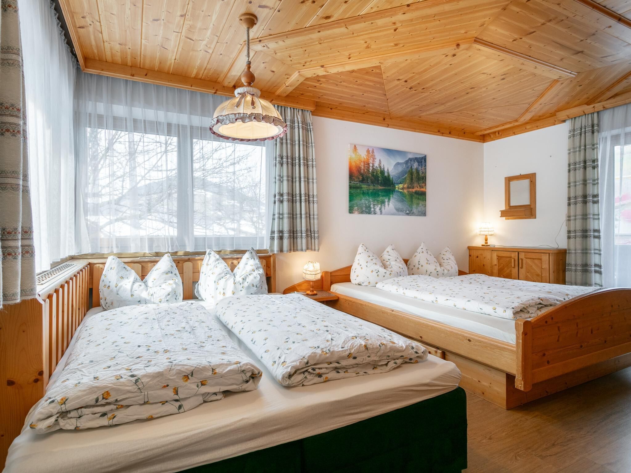 A room with two beds, wooden headboards, a wooden ceiling, a painting of a landscape, and a window with curtains.