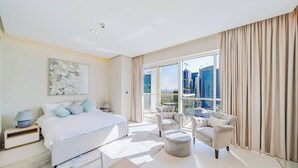 Quarto de hotel moderno com uma cama grande, área de estar com duas poltronas e um banquinho redondo, janela ampla com vista para a cidade e teto com iluminação embutida.