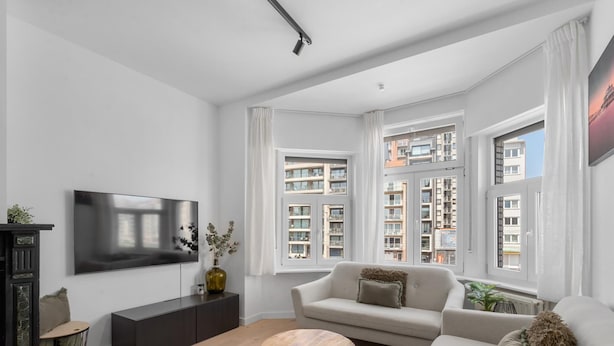 A modern living room with a flat-screen TV, a sofa, a coffee table, and large windows offering a view of the cityscape. - Renovated apartment for family (Blankenberge)