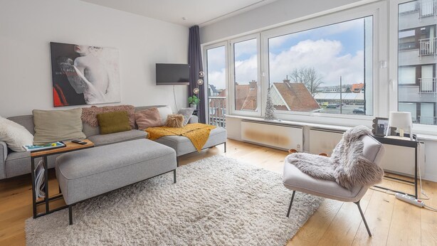 Ein modernes Wohnzimmer mit einem grauen Sofa, einem weißen Sessel und einem großen Fenster mit Blick auf die Stadt.