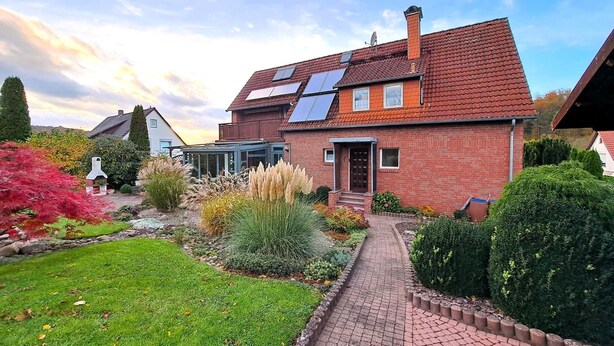 A house with a brick exterior, a tiled roof, and a garden with various plants and a pathway. - Holiday apartment with balcony in Weserbergland (Emmerthal OT Welsede)