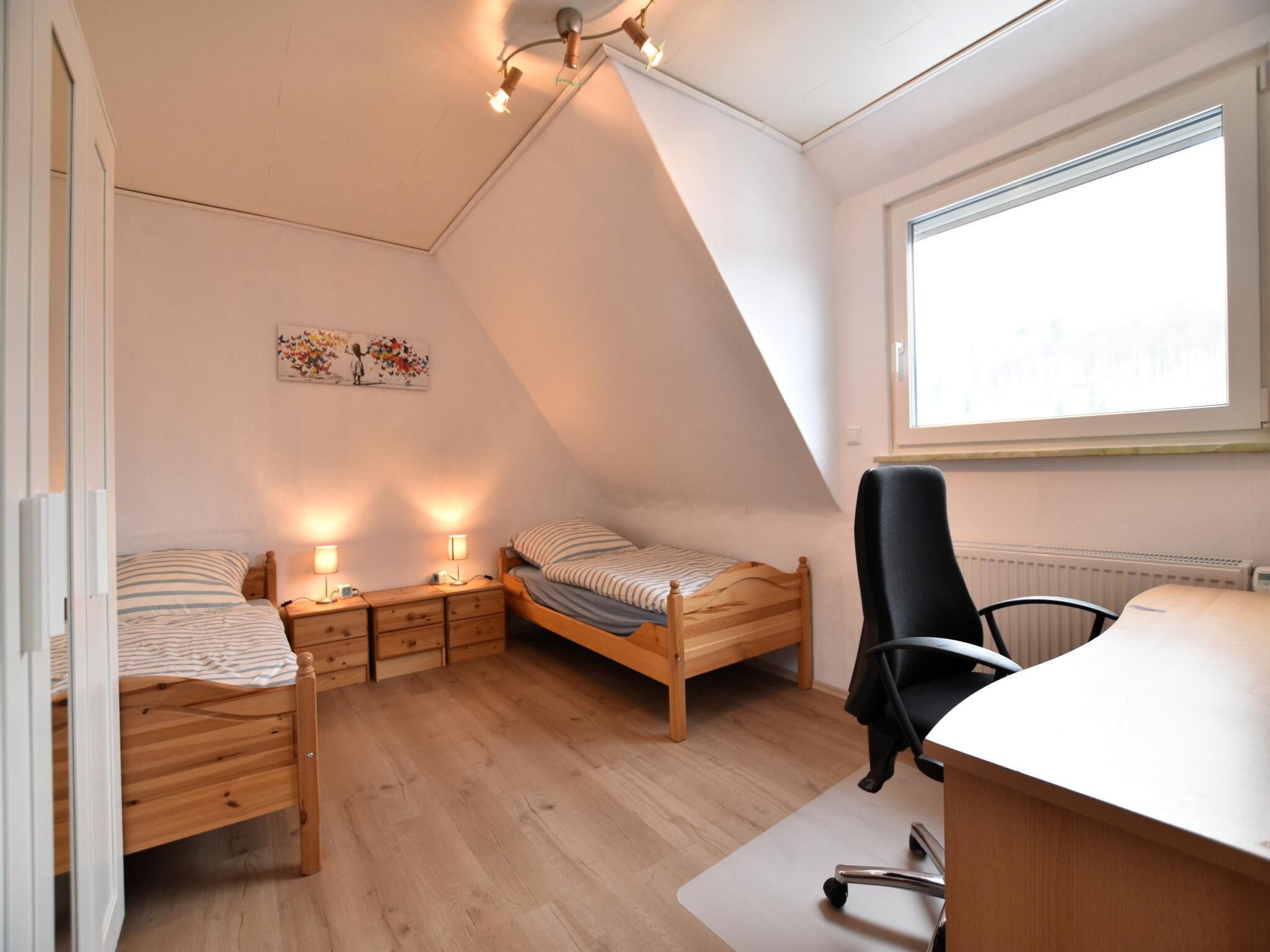 A small, well-lit bedroom with two single beds, a desk, and a chair.