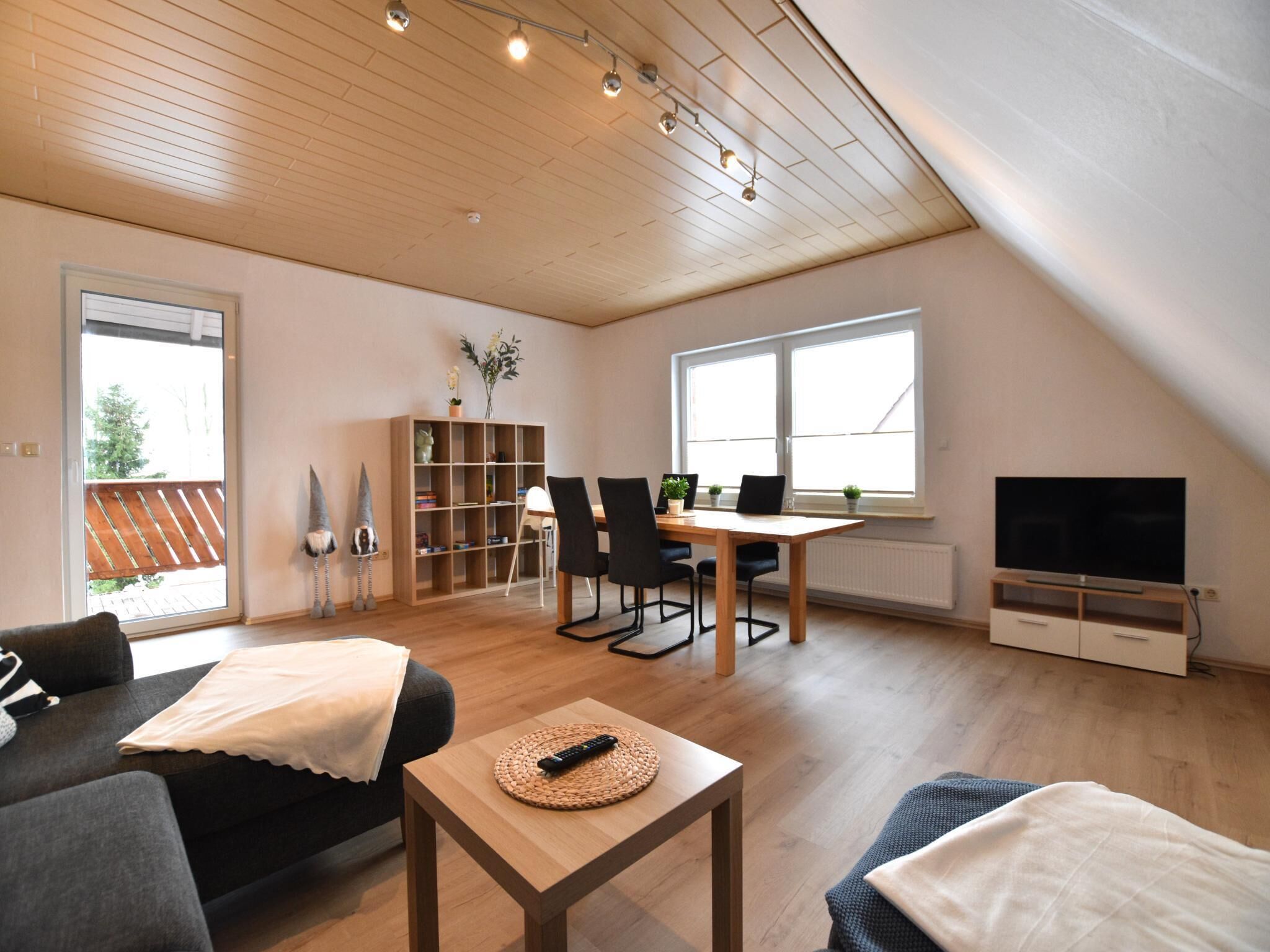 A spacious living room with a wooden ceiling, a dining area with a table and chairs, a bookshelf, a TV, and a sofa.