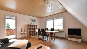 A spacious living room with a wooden ceiling, a dining area with a table and chairs, a bookshelf, a TV, and a sofa. - Holiday apartment with balcony in Weserbergland (Emmerthal OT Welsede)