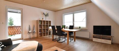 A spacious living room with a wooden ceiling, a dining area with a table and chairs, a bookshelf, a TV, and a sofa.