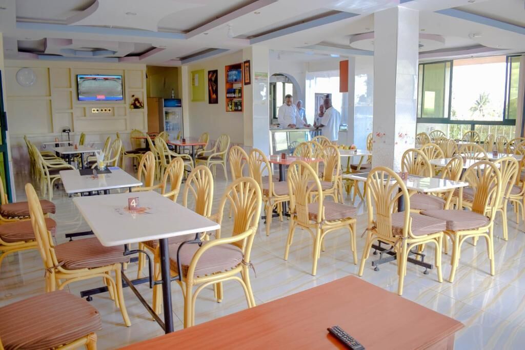 Un restaurant avec des chaises en osier et des tables blanches, un téléviseur accroché au mur et un espace cuisine à l’arrière-plan.