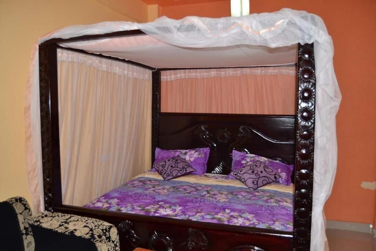 A four-poster bed with a canopy, purple floral bedspread, and matching pillows.