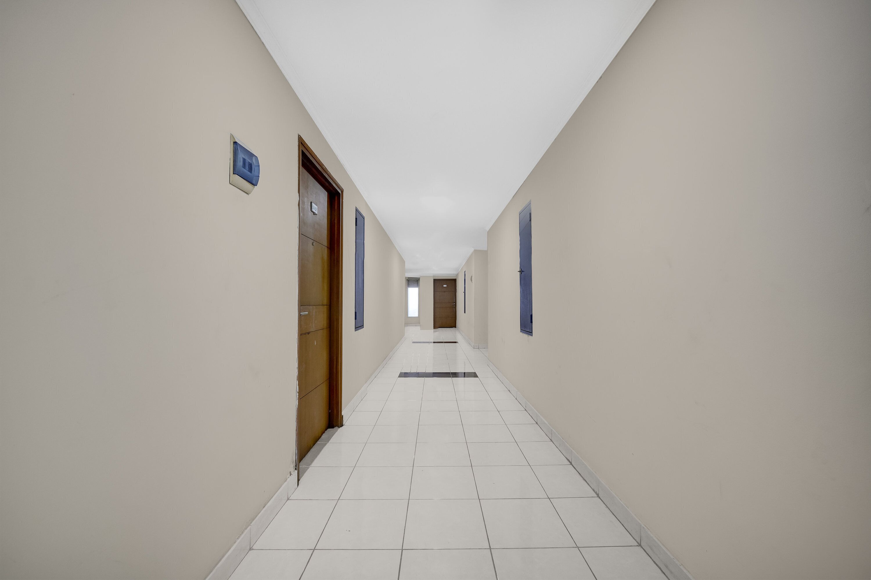 A long, narrow hallway with white tiled floors and beige walls. There are doors on both sides, some with blue frames.