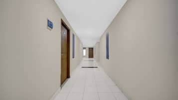 A long, narrow hallway with white tiled floors and beige walls. There are doors on both sides, some with blue frames.