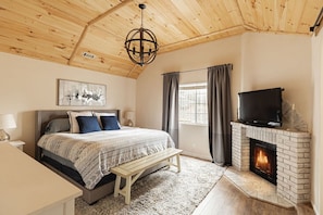 A bedroom with a bed, bedside tables, a bench, a fireplace, and a television. - Elevated Escape | Bocce, Frisbee Glf, Walk to Town (Big Bear Lake)