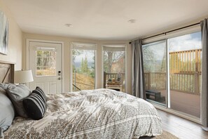 A bedroom with a large bed, a door leading to a balcony, and a view of a wooden deck. - Elevated Escape | Bocce, Frisbee Glf, Walk to Town (Big Bear Lake)