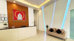 A reception area with a wooden reception desk, a large framed artwork, a beige sofa, and a potted plant. - Foura Suites Makati (Makati)