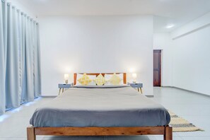 Un dormitorio moderno con un cabecero de madera, una cama grande con ropa de cama gris, dos mesitas de noche con lámparas y un ventanal con cortinas.