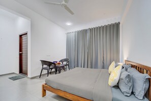 Un dormitorio con cama, escritorio, dos sillas, ventilador de techo y ventana con cortinas.