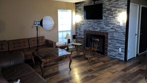 A living room with a brown leather sofa, a glass-top coffee table, a fireplace with a stone wall, and a flat-screen TV mounted above the fireplace. - Quiet ground floor 2 bedroom- unwind in private. (Garland)