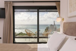 Quarto com uma janela ampla voltada para o oceano, uma cama com roupa de cama branca e duas cadeiras de vime em uma varanda externa.