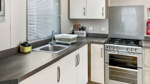 Une cuisine moderne équipée d’une cuisinière, d’un four et d’un évier en acier inoxydable. On y trouve des armoires blanches, une fenêtre avec des stores et un porte-ustensiles sur le plan de travail.