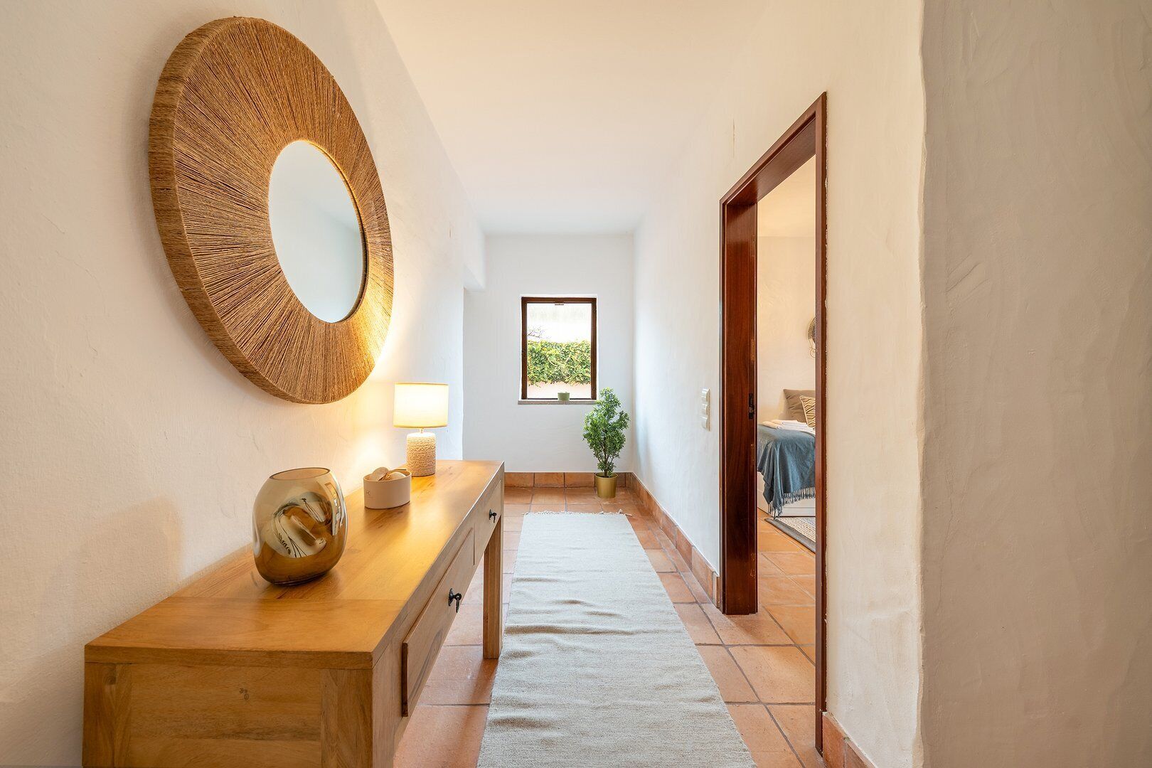 Un couloir avec une console en bois, un miroir rond, une plante en pot et une fenêtre donnant sur un paysage verdoyant.