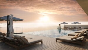 Une piscine à débordement située sur le toit, avec des chaises longues et des parasols, offrant une vue imprenable sur l’océan au coucher du soleil.