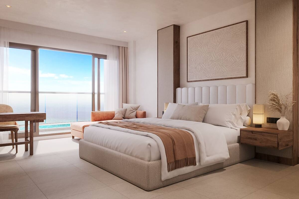Une chambre moderne avec un grand lit, une table de chevet avec une lampe et une vue sur l’océan grâce à de grandes fenêtres.