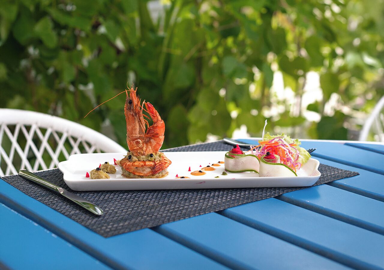 Une table à manger avec une assiette blanche contenant des aliments, notamment une crevette et une salade, est placée sur une table bleue. Une chaise blanche se trouve à l'arrière-plan.