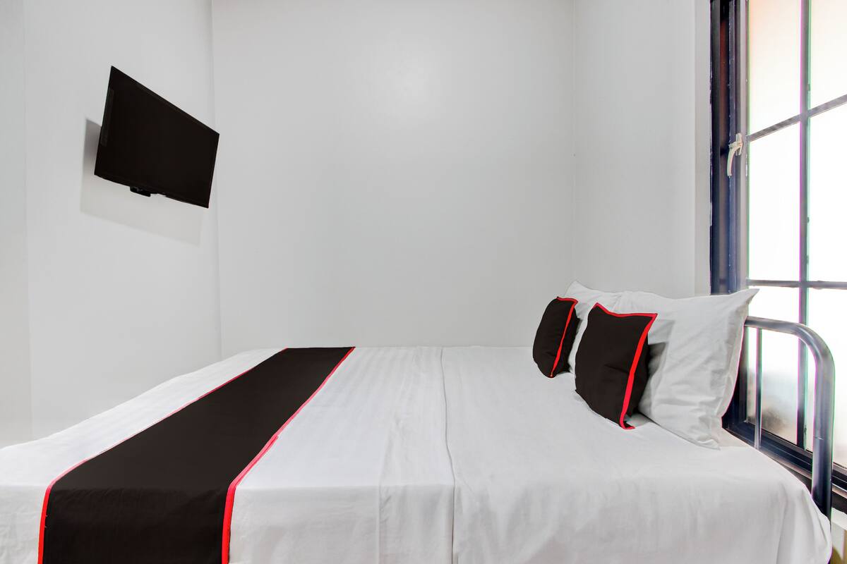 Una cama individual con colcha blanco y negro, un televisor de pantalla plana en la pared, y una ventana con marco de metal.