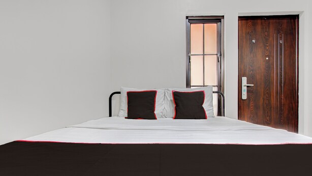 Un lit recouvert d’une couvre-lit noire et blanche, deux oreillers noirs bordés de rouge, une porte en bois et une fenêtre donnant sur l’extérieur.