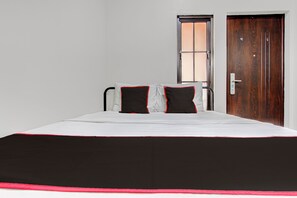 Un lit recouvert d’une couvre-lit noire et blanche, deux oreillers noirs bordés de rouge, une porte en bois et une fenêtre donnant sur l’extérieur.