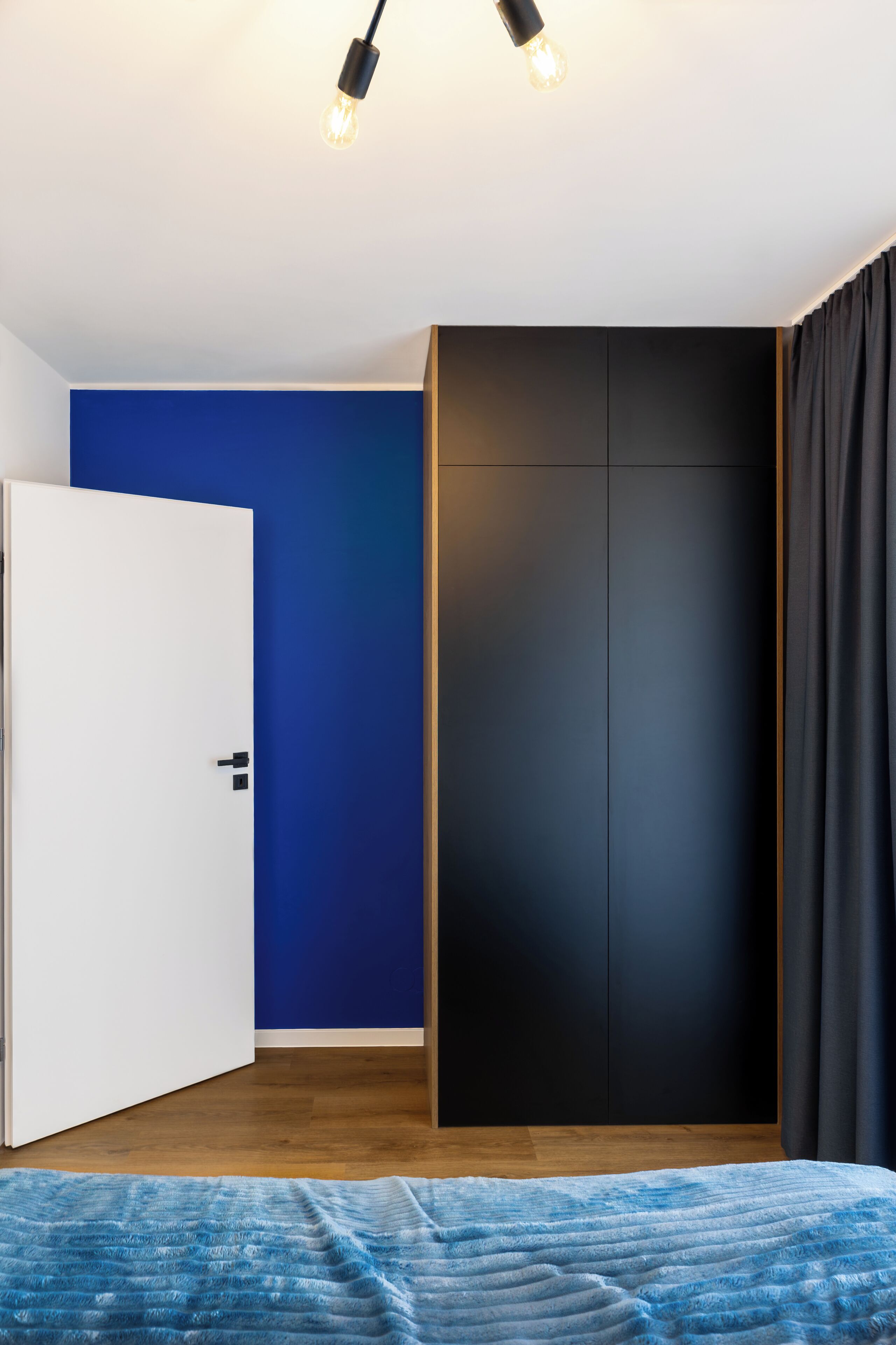 A bedroom with a white door, a blue wall, a black wardrobe, and a bed with a blue blanket.