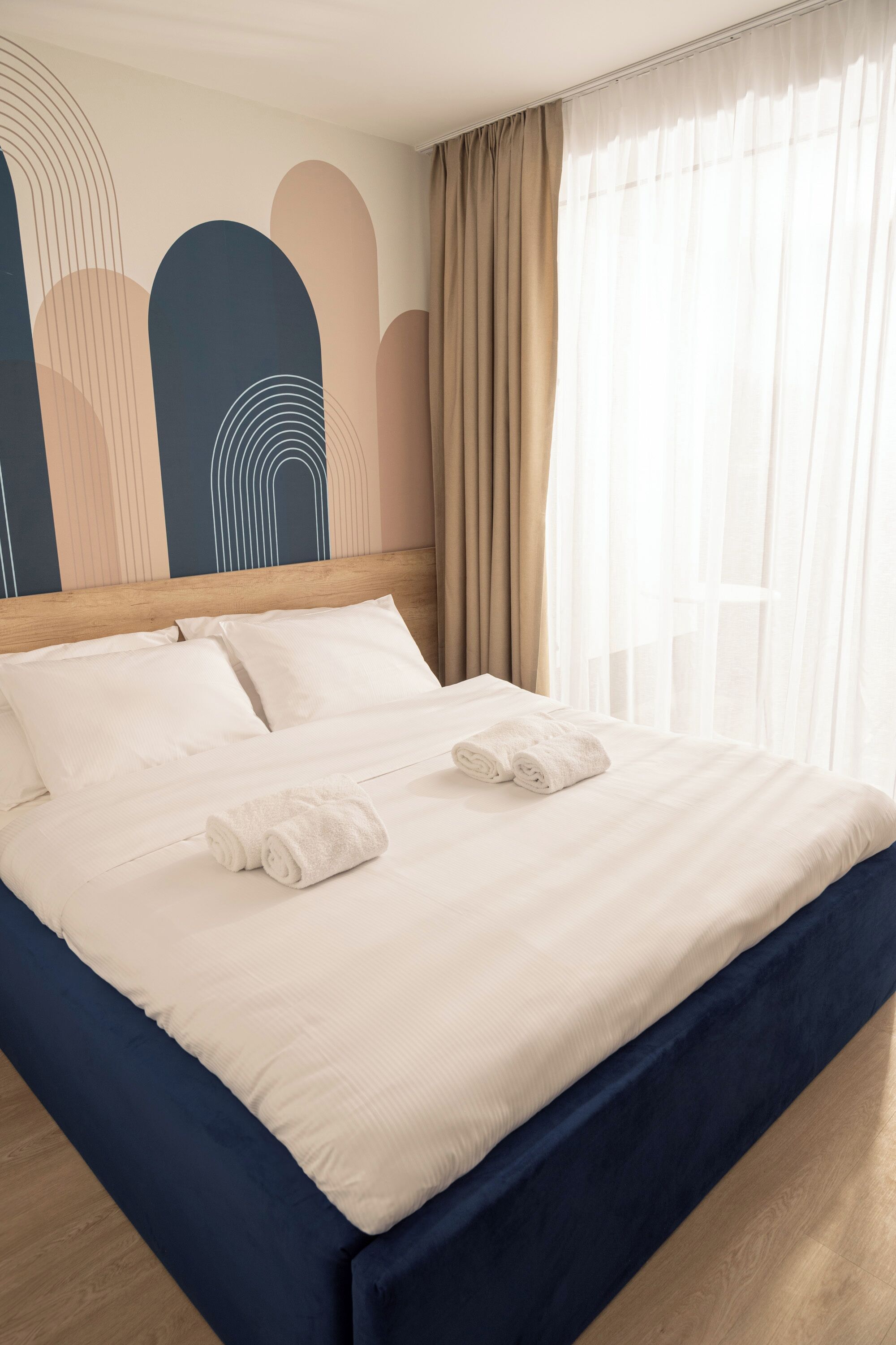 Una cama bien hecha con sábanas blancas y dos toallas enrolladas, un cabecero de madera y una pared con arte abstracto.