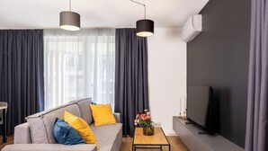 A modern living room with a grey sofa, yellow and blue cushions, a wooden coffee table, a flat-screen TV, and a large window with dark curtains. - Baltic Season Spa Apartments (Grzybowo)