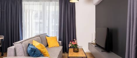 Una sala de estar moderna con un sofá gris, cojines amarillos y azules, una mesa de centro de madera, un televisor de pantalla plana y un ventanal con cortinas oscuras.