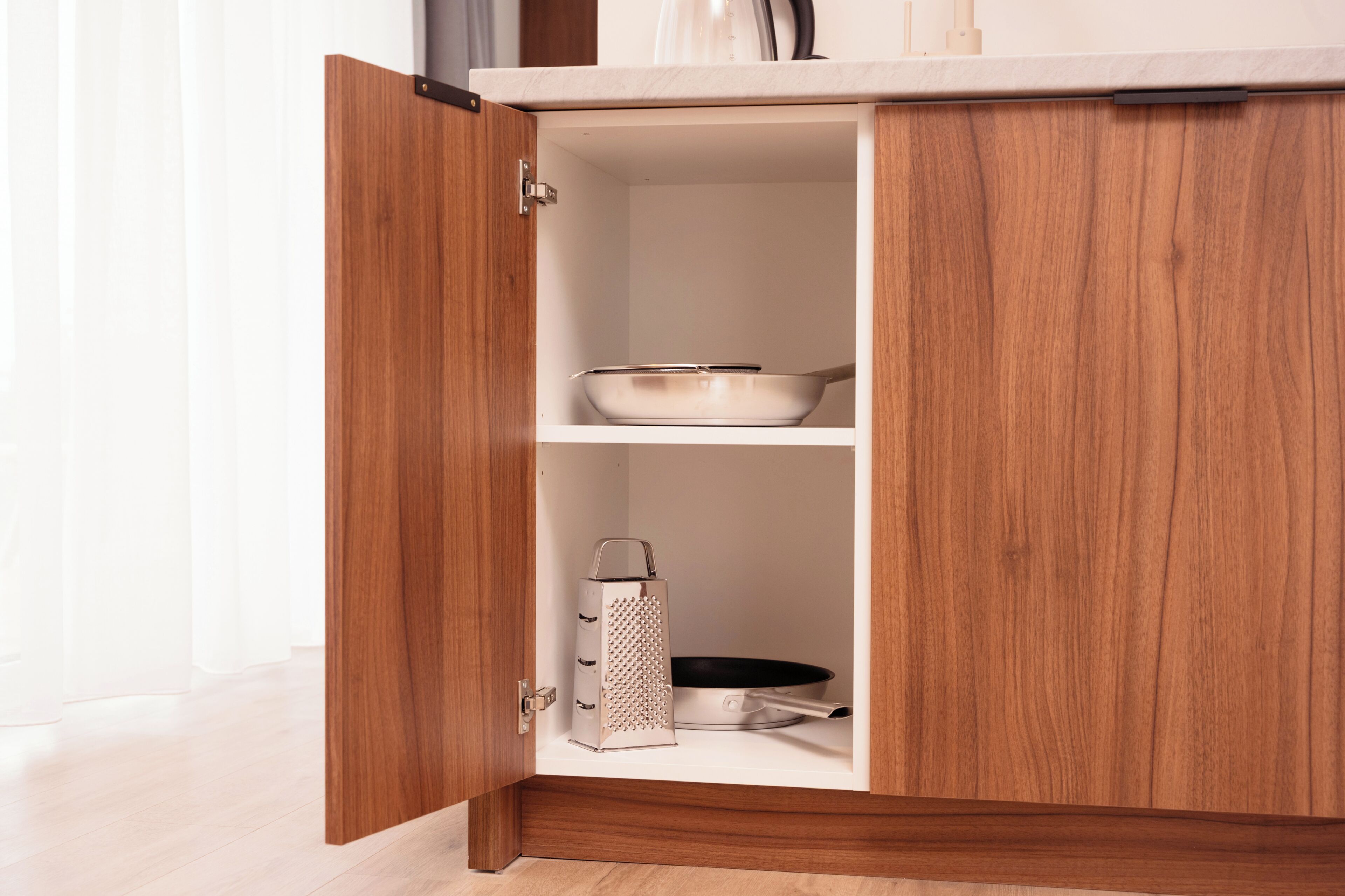 A wooden cabinet with a white interior, containing a grater and a frying pan.