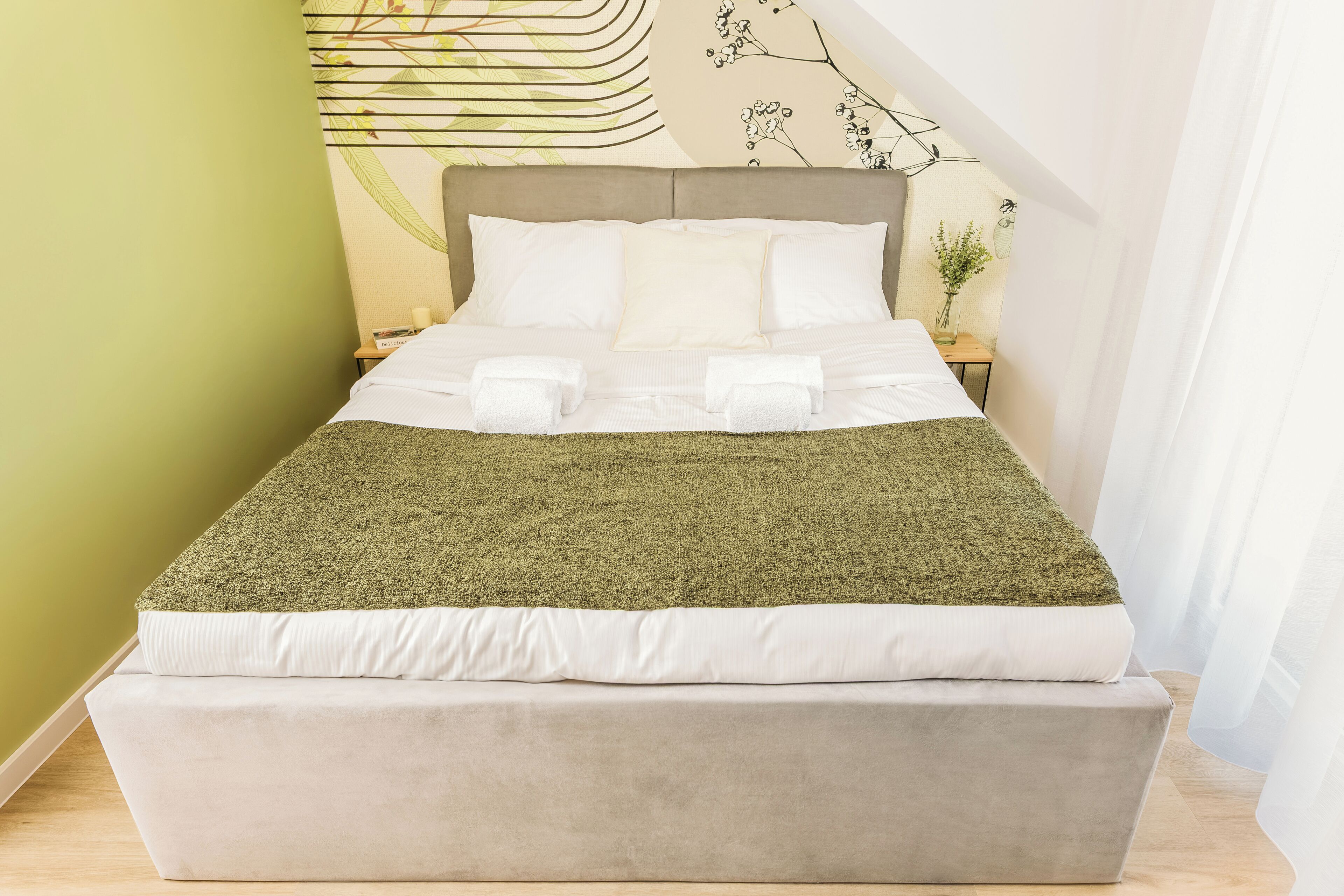 A bed with a green throw, white bedding, and a headboard with a wall art design.