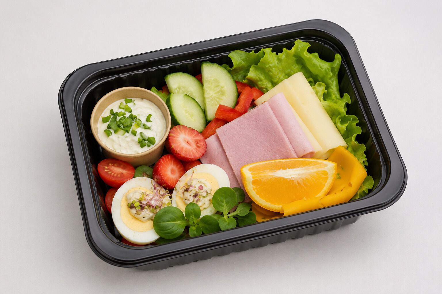 Comida en un recipiente negro con lechuga, rodajas de pepino, tomates, fresas, huevos cocidos, jamón, queso y una naranja.