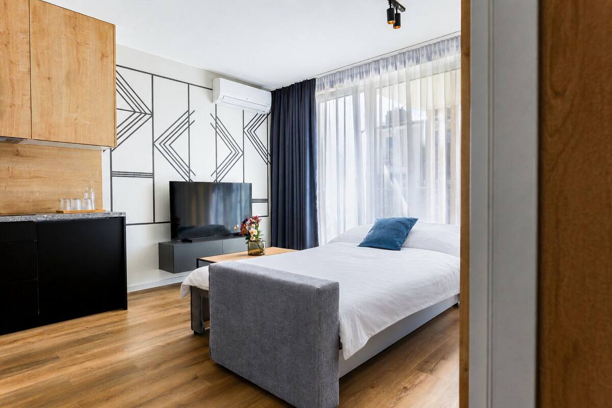 Ein modernes Hotelzimmer mit Holzboden, einem Bett mit grauem Kopfteil, einem Fernseher auf einem schwarzen Standfuß, einem kleinen Tisch mit Blumen in einer Vase und einem großen Fenster mit transparenten Vorhängen.