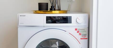 A white Sharp washing machine with a control panel, a small plant on top, and a power outlet nearby.