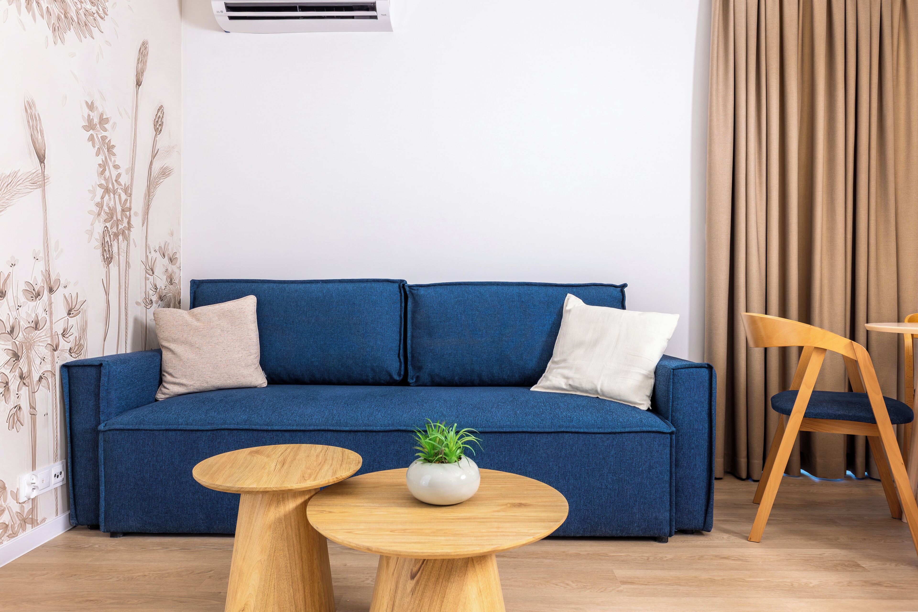 A blue sofa with two pillows, a small white plant on a wooden table, and a wooden chair.