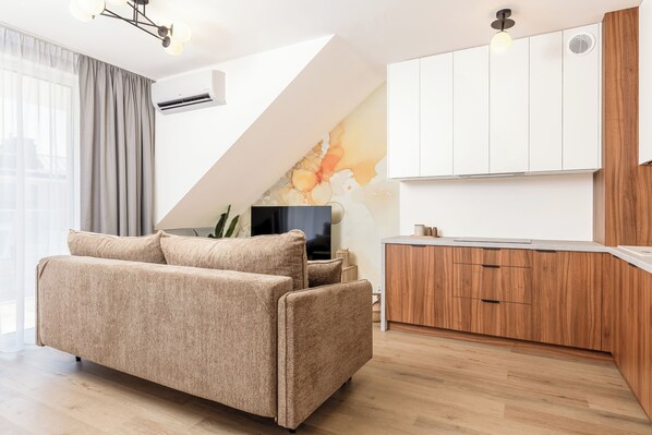 A modern living room with a sofa, a flat-screen TV, and a kitchen area with wooden cabinets and a white countertop.
