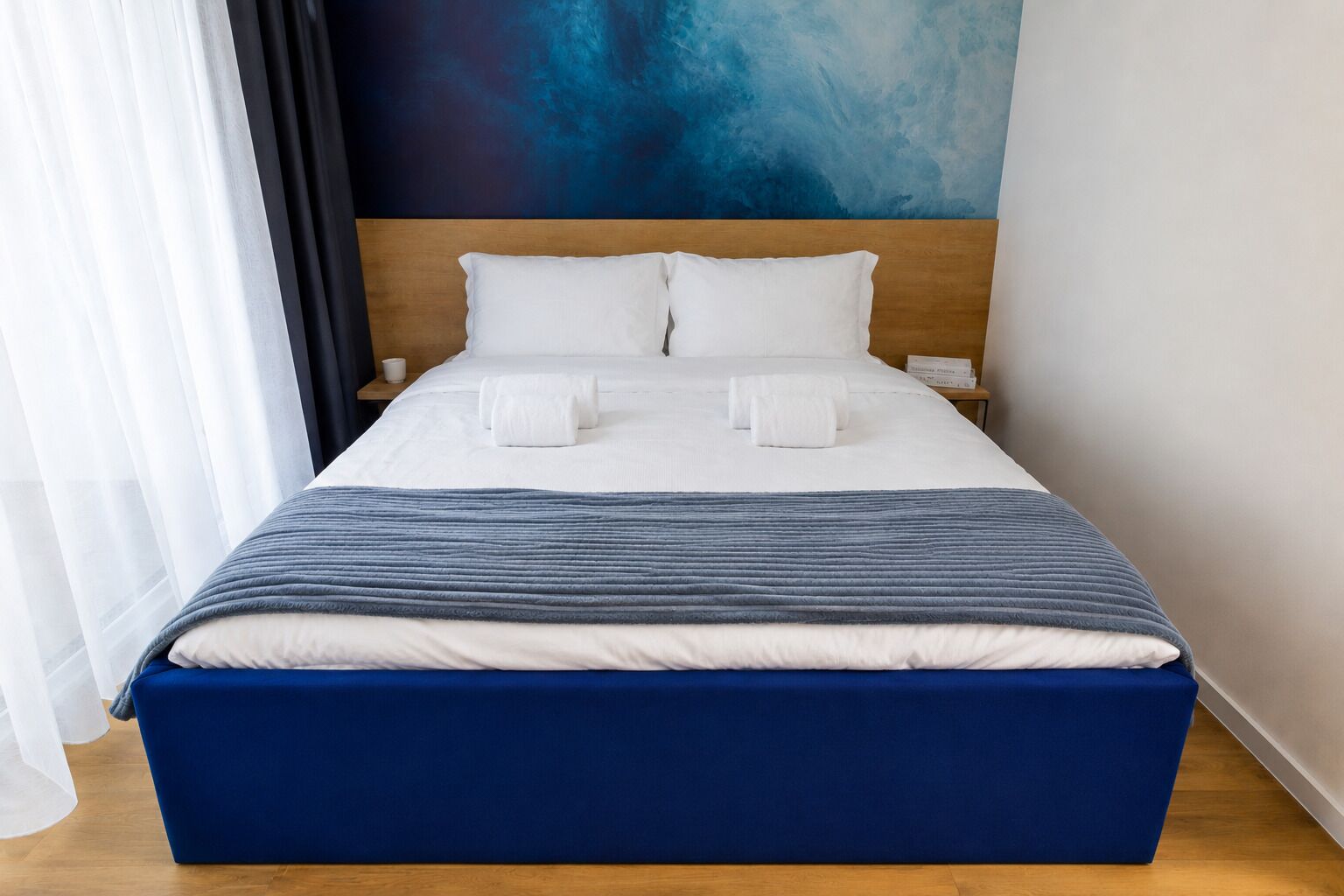 A neatly made bed with white linens and a blue accent, a wooden headboard, and a blue abstract painting on the wall.