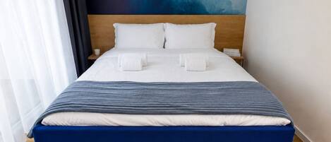 A neatly made bed with white linens and a blue accent, a wooden headboard, and a blue abstract painting on the wall.