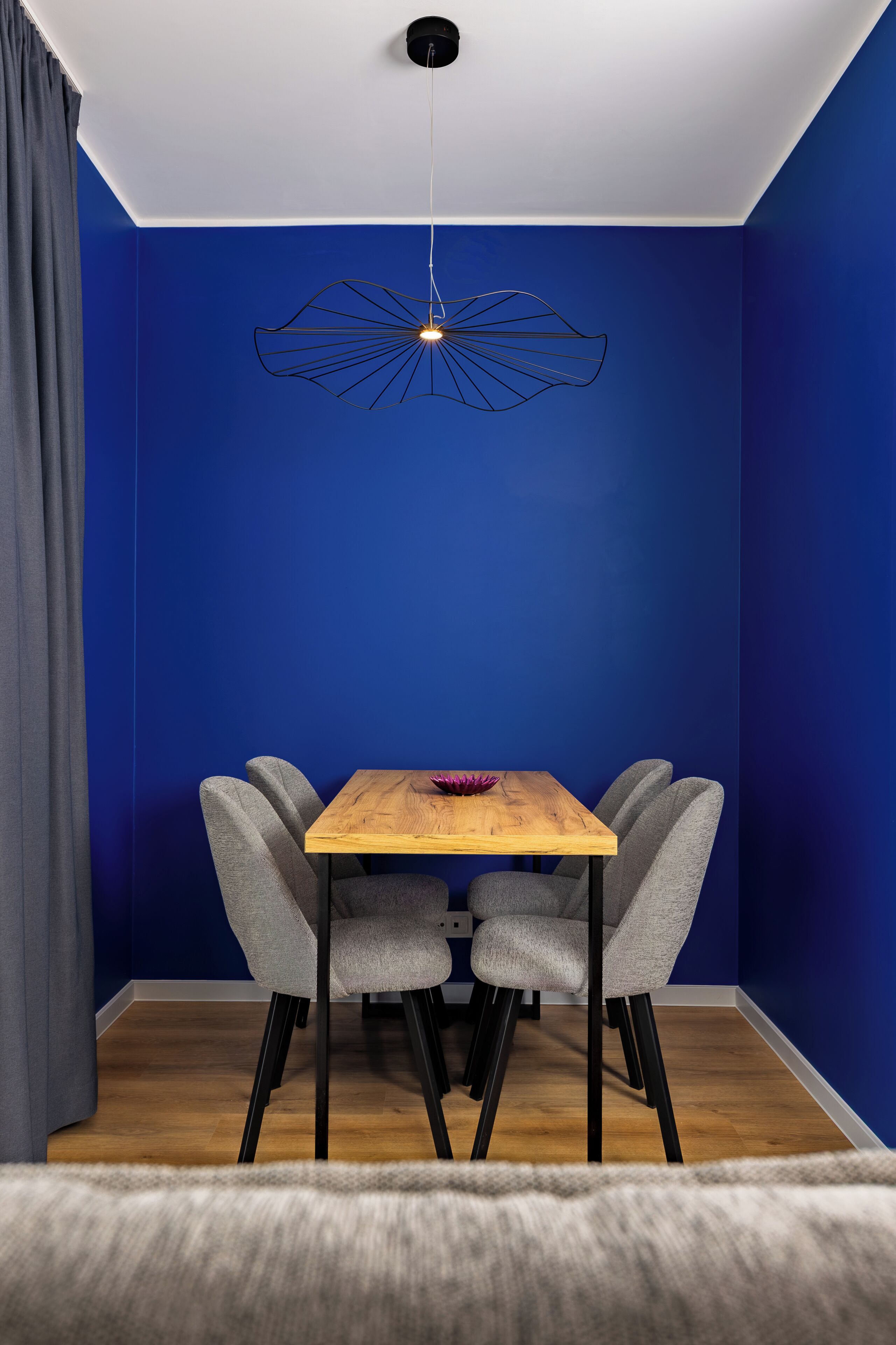 A room with a blue wall, a wooden table, four grey chairs, a hanging light fixture, and a grey curtain.