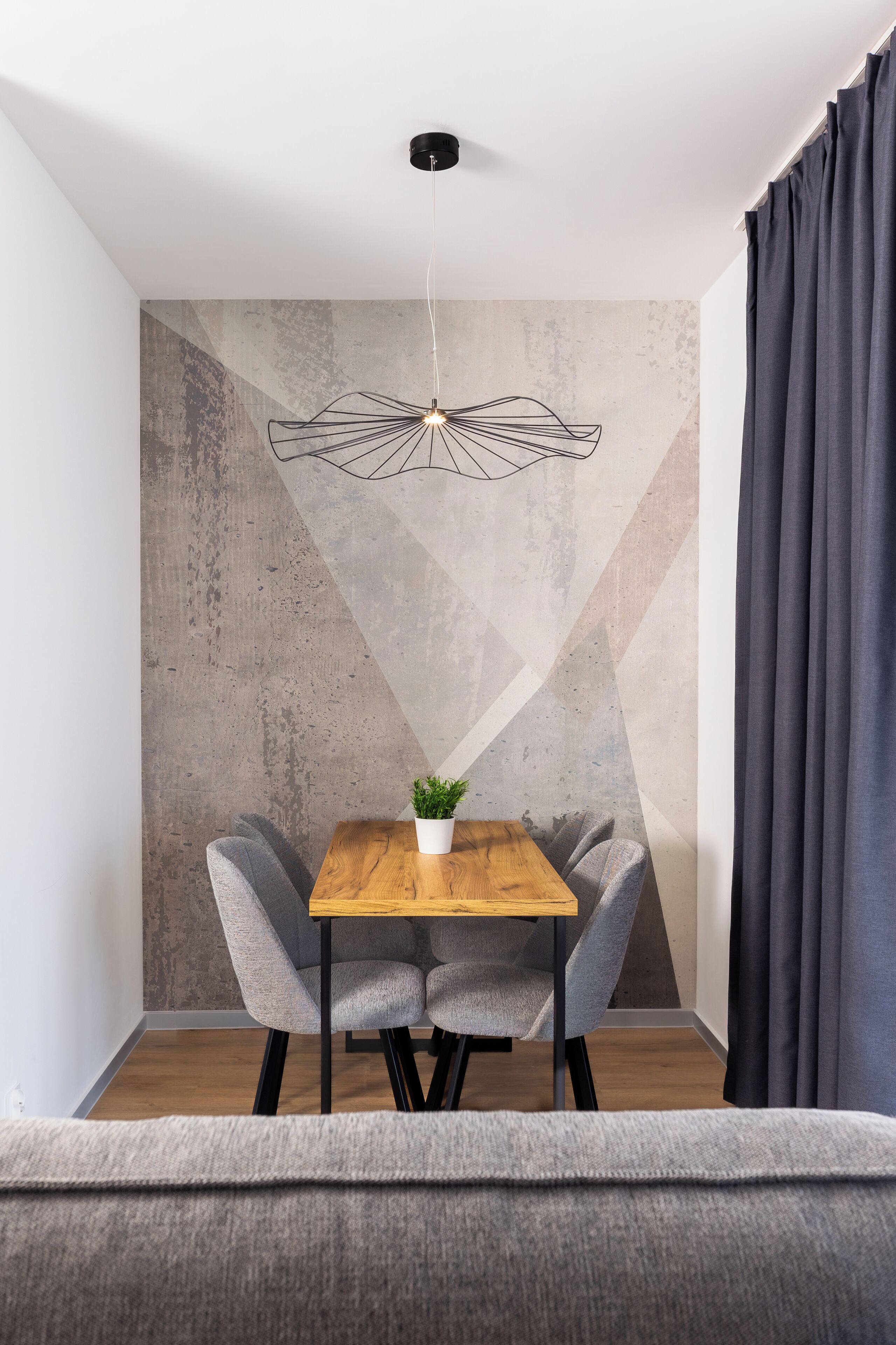 Un comedor con mesa de madera, dos sillas grises, una planta en maceta y una lámpara colgante moderna.