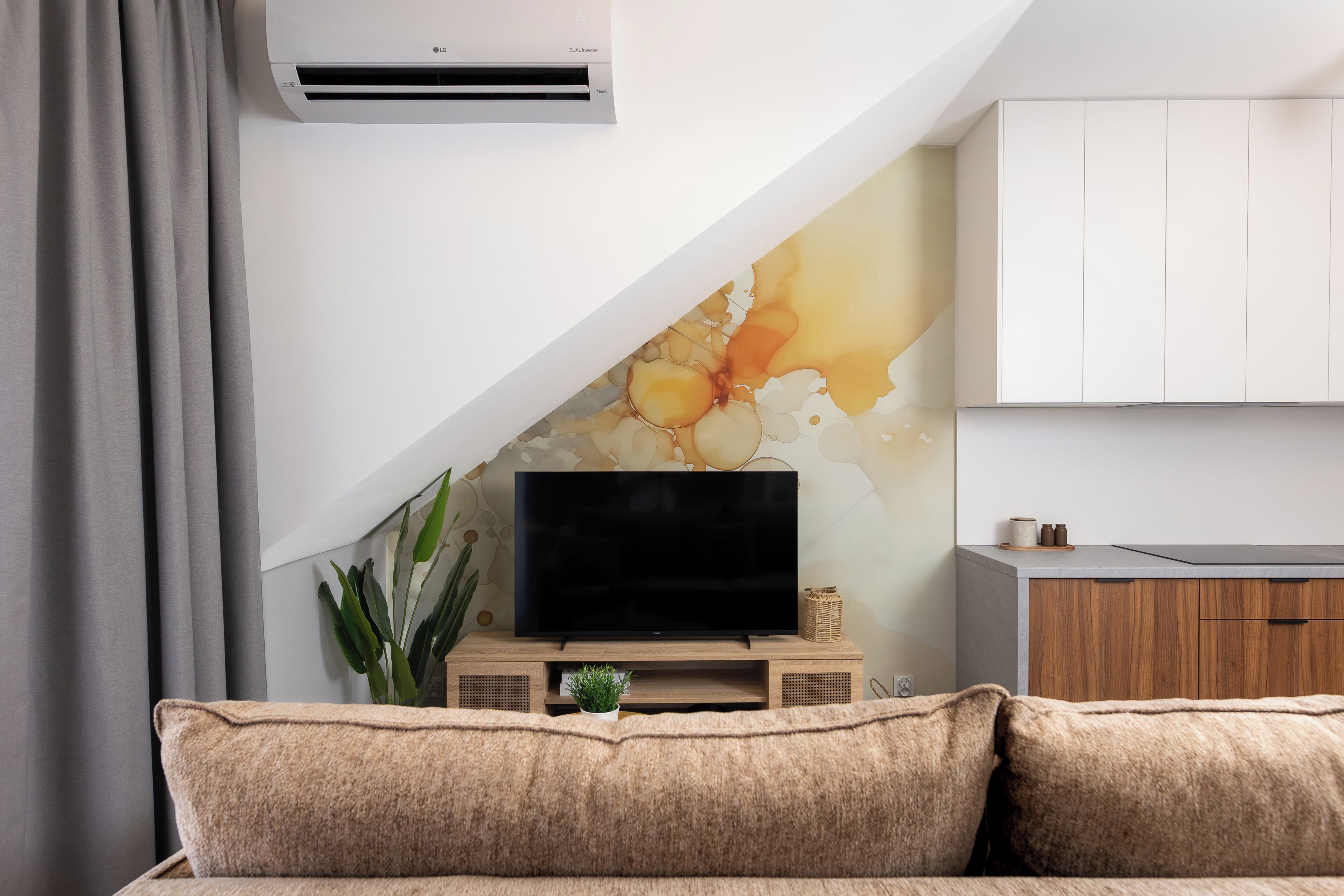 A living room with a flat-screen TV on a wooden stand, a grey couch, a potted plant, and a wall-mounted air conditioner.