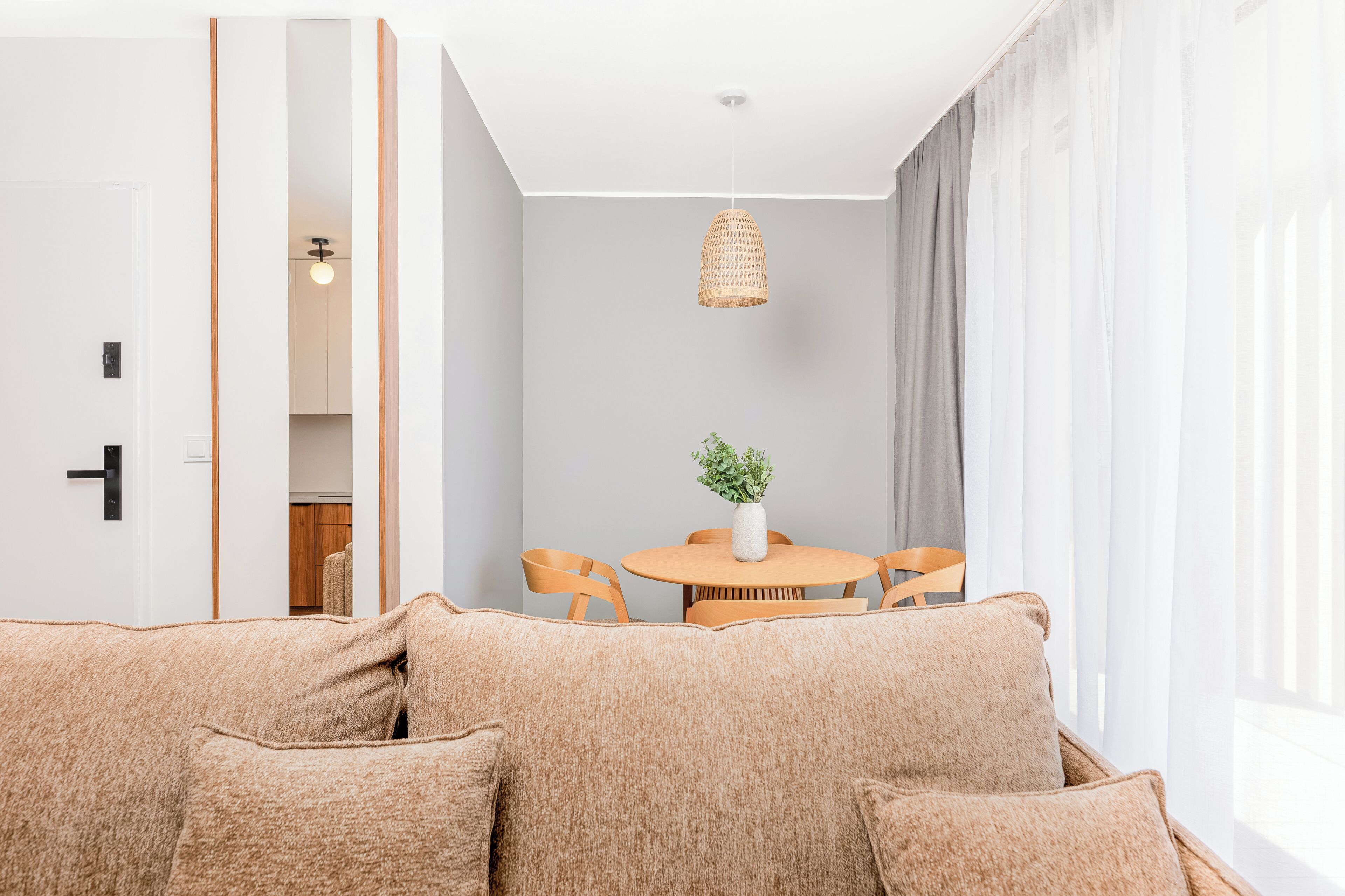 Ett modernt vardagsrum med en beige soffa, ett runt träbord med stolar, en hängande lampa och utsikt över ett kök genom en öppen dörr.