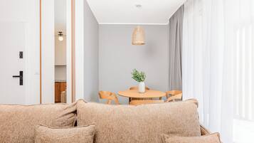 Ett modernt vardagsrum med en beige soffa, ett runt träbord med stolar, en hängande lampa och utsikt över ett kök genom en öppen dörr.