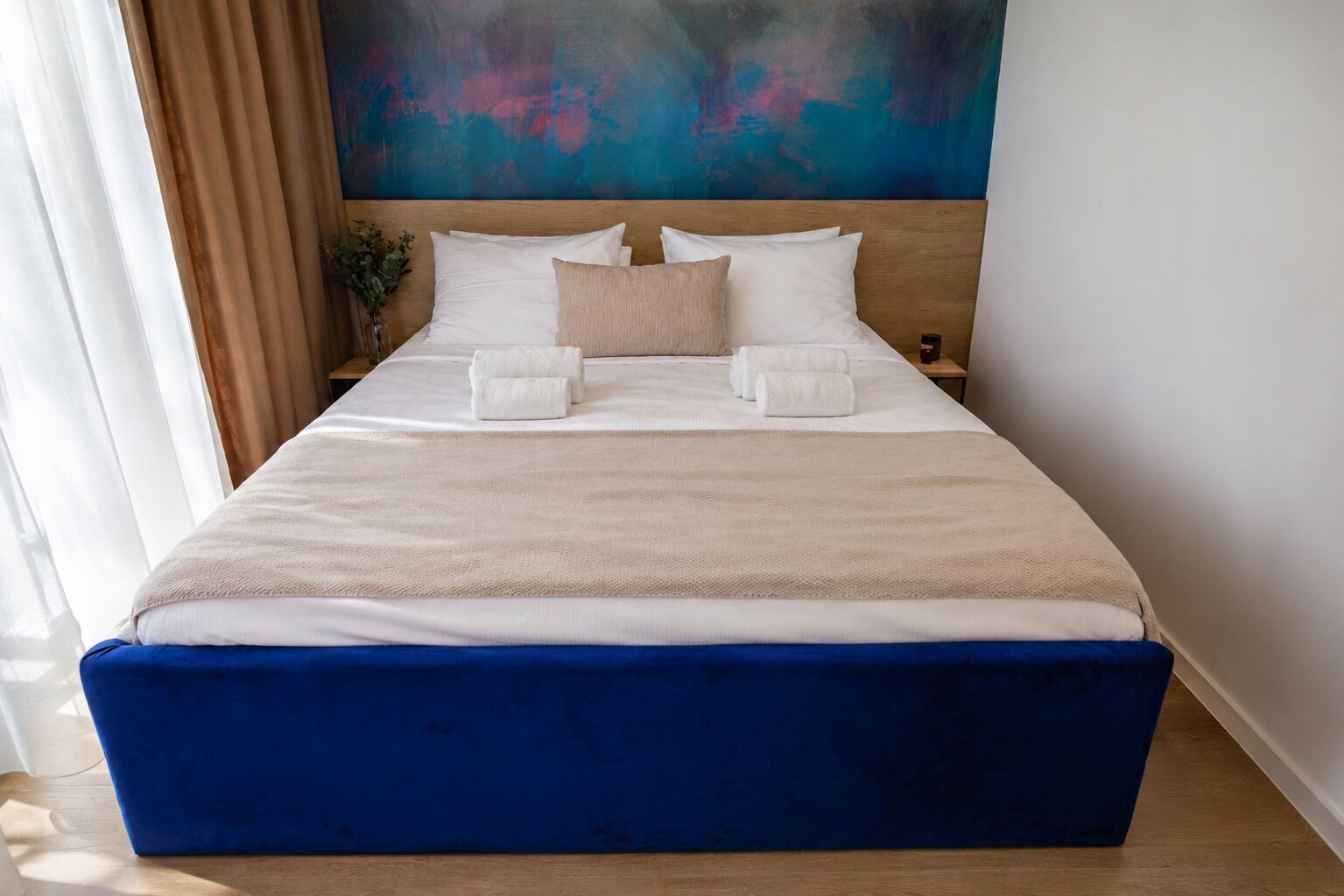 Een groot bed met wit en beige beddengoed, een blauw hoofdeinde en een schilderij erboven.