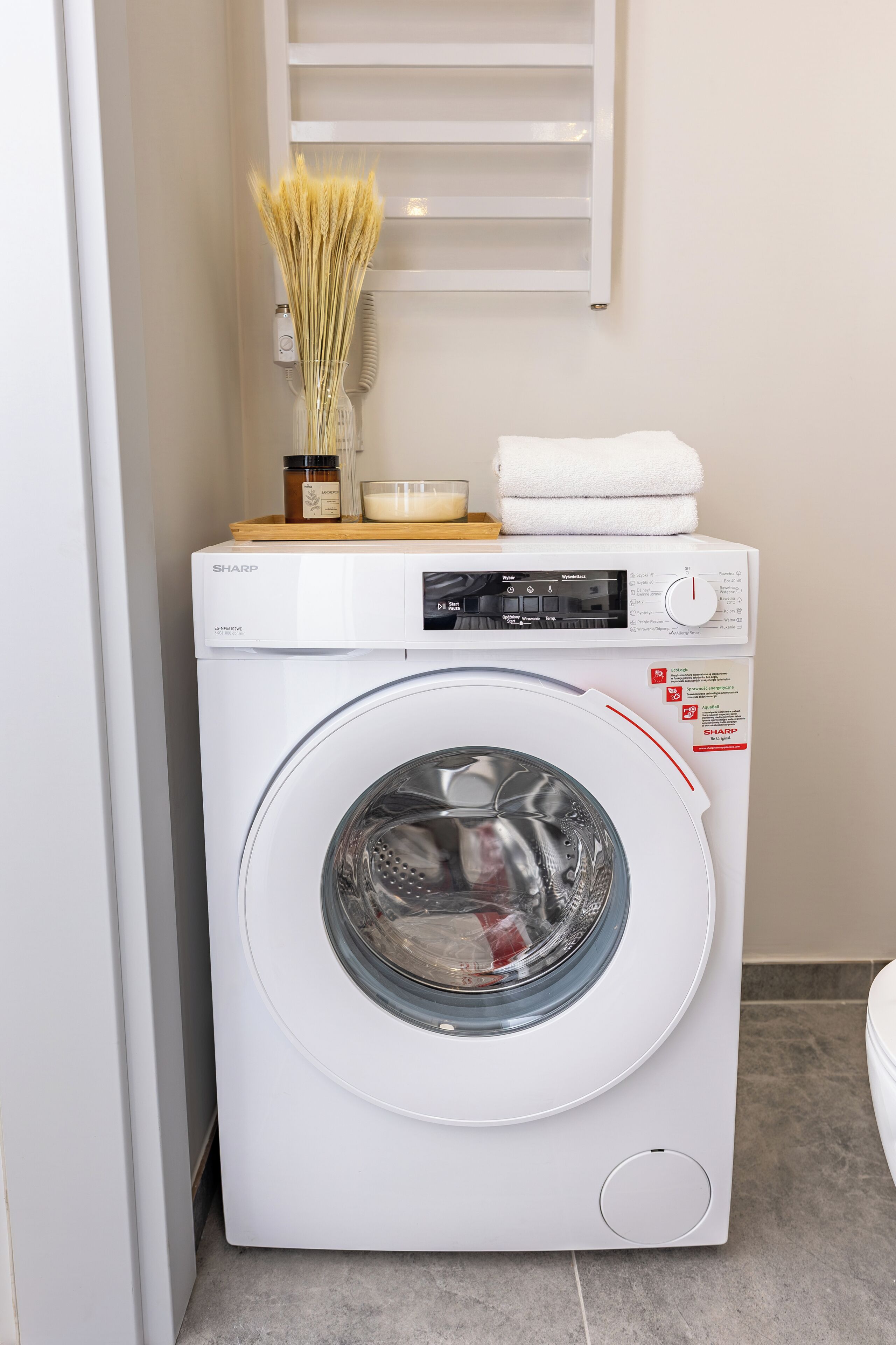 A front-loading washing machine with a control panel, a small shelf with a candle, a jar, and a vase with dried plants.