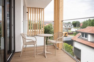 Un balcón con una mesa y dos sillas, listones de madera y vistas a casas y follaje.