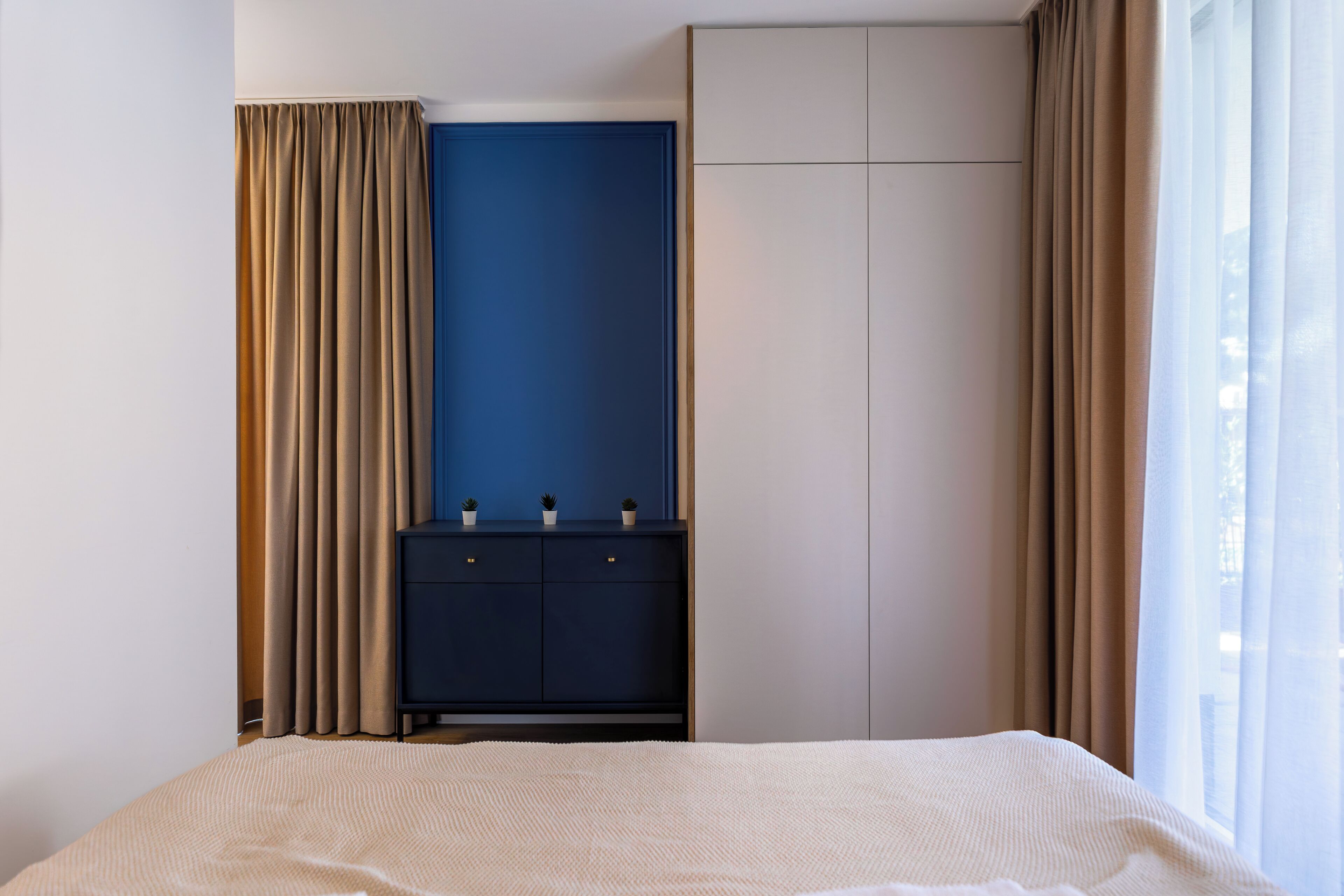 A bedroom with a bed, a dark blue wall, a wardrobe, and a window with curtains.